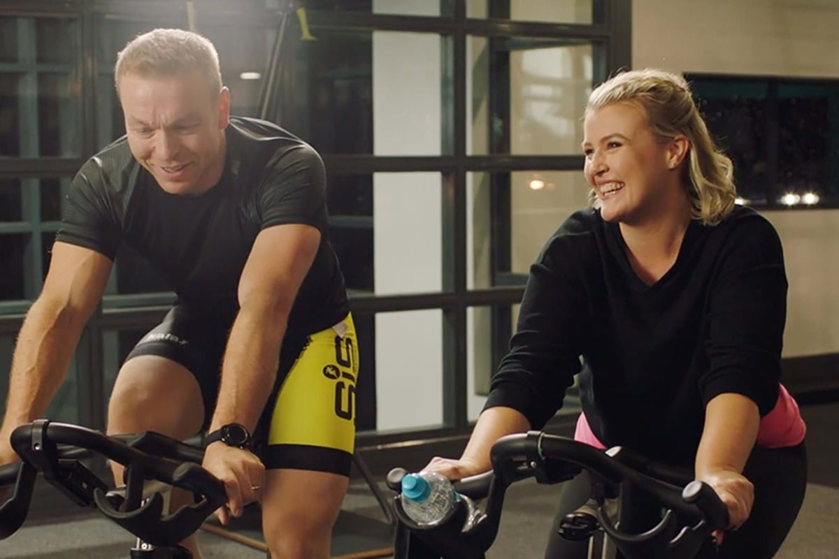 Man and woman cycling indoors, woman smiling and holding a water bottle on bike handlebar