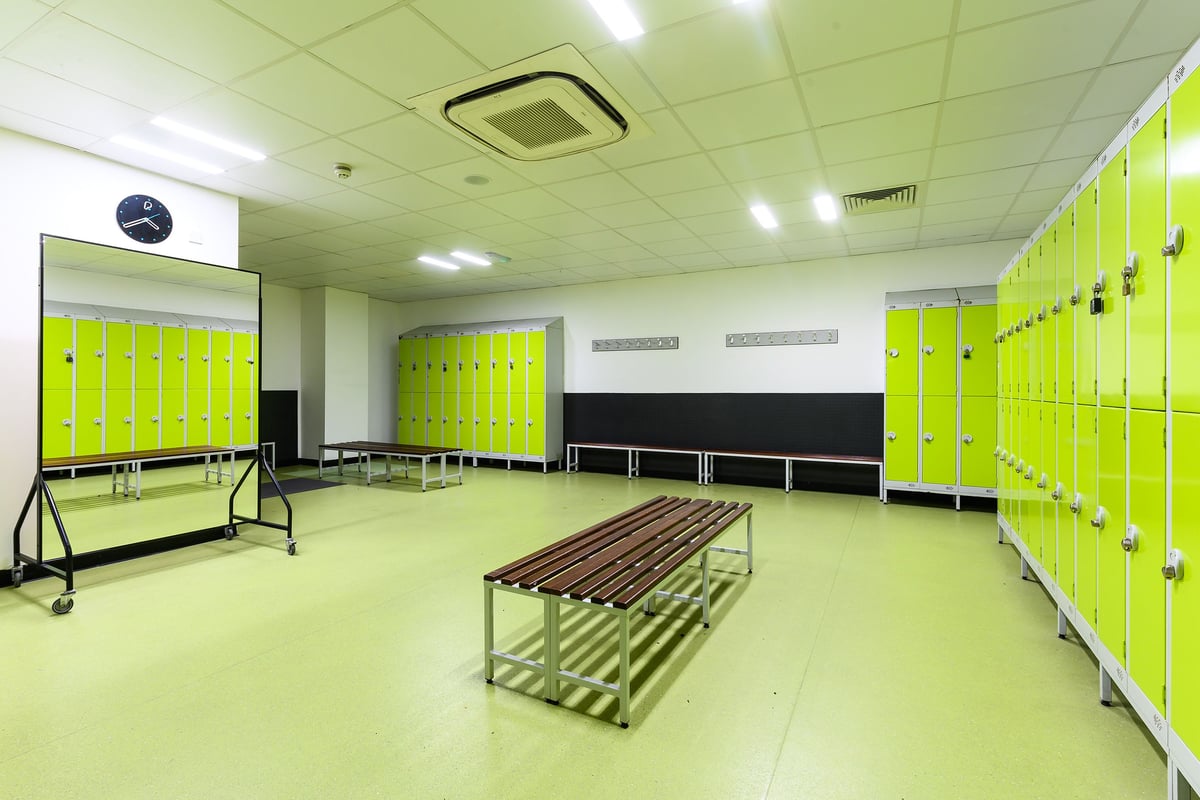 PureGym Bromborough: Rows of bright green lockers, wooden benches, and an equipment trolley in a changing room.