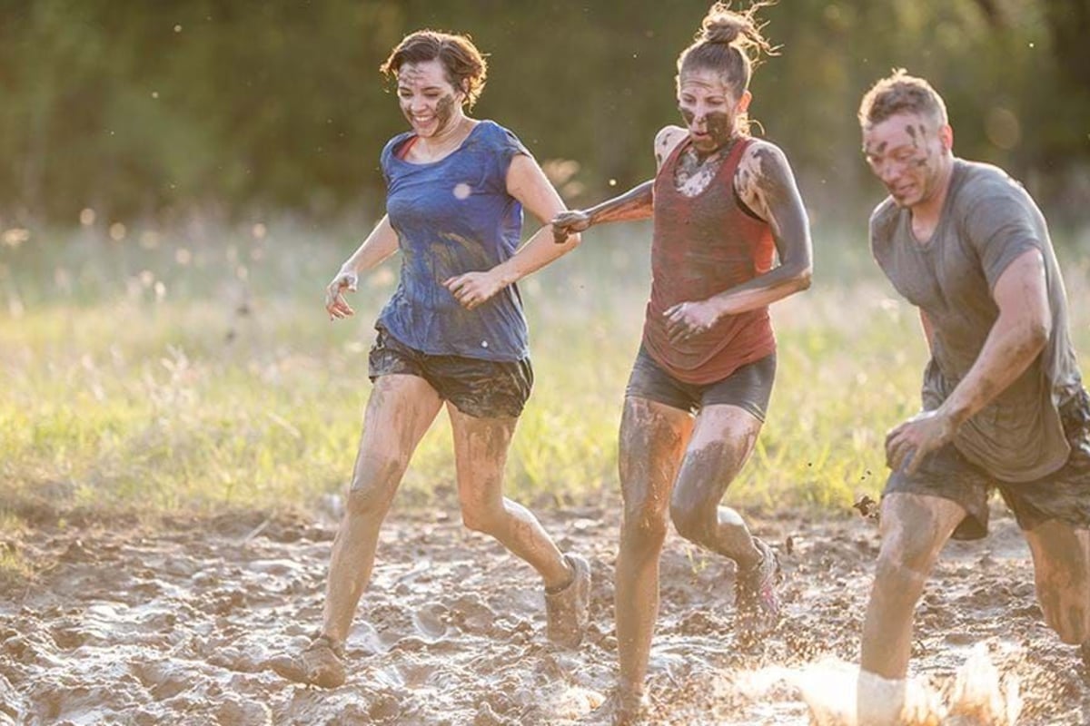 Three people covered in mud running and holding hands in a muddy field at sunset