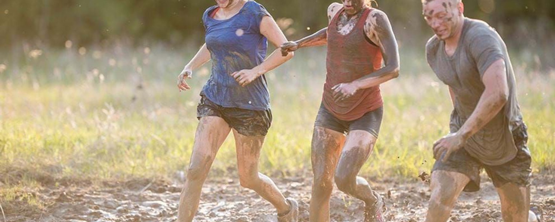Three people covered in mud running and holding hands in a muddy field at sunset