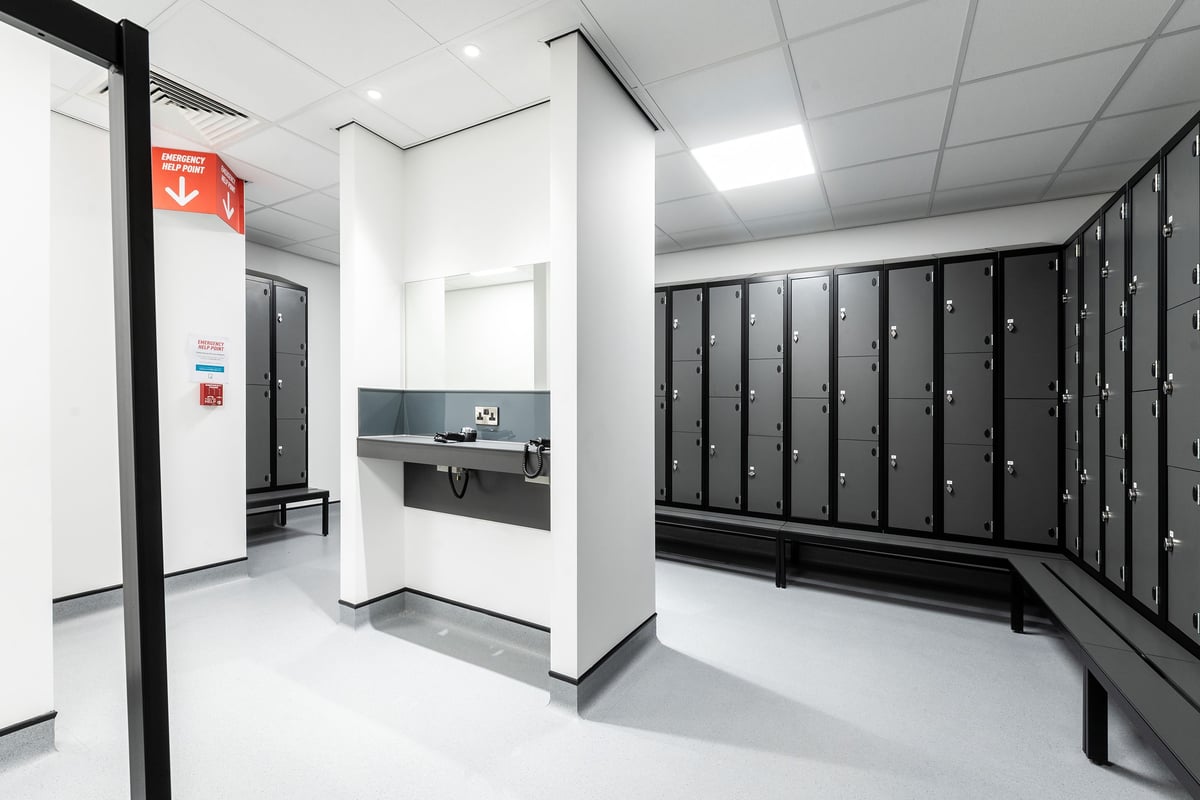 PureGym Cardiff Western Avenue: Rows of lockers, a counter, and a fire extinguisher in a changing room setting.