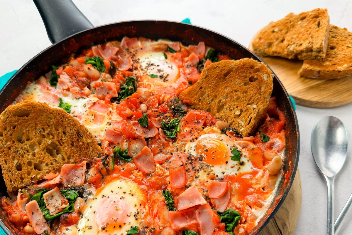 Frying pan with baked eggs, beans, tomatoes, spinach and bacon beside toasted bread