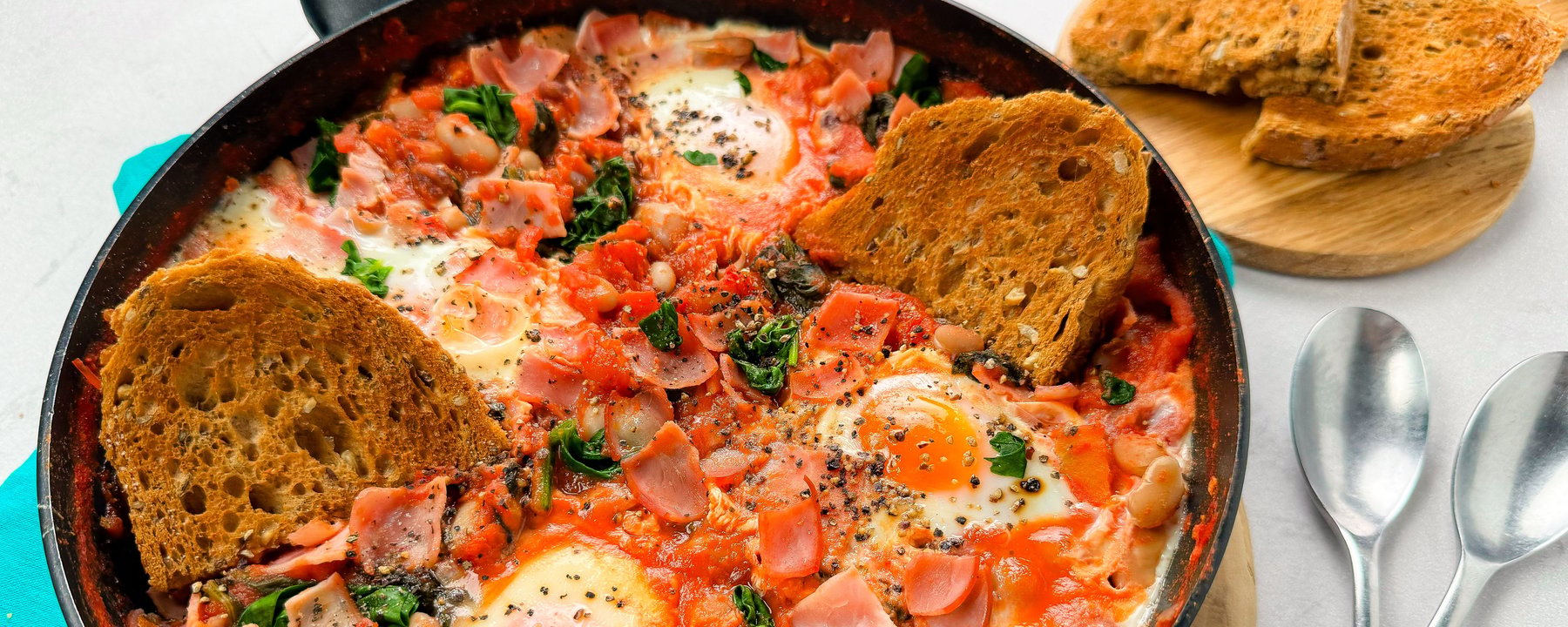 Frying pan with baked eggs, beans, tomatoes, spinach and bacon beside toasted bread