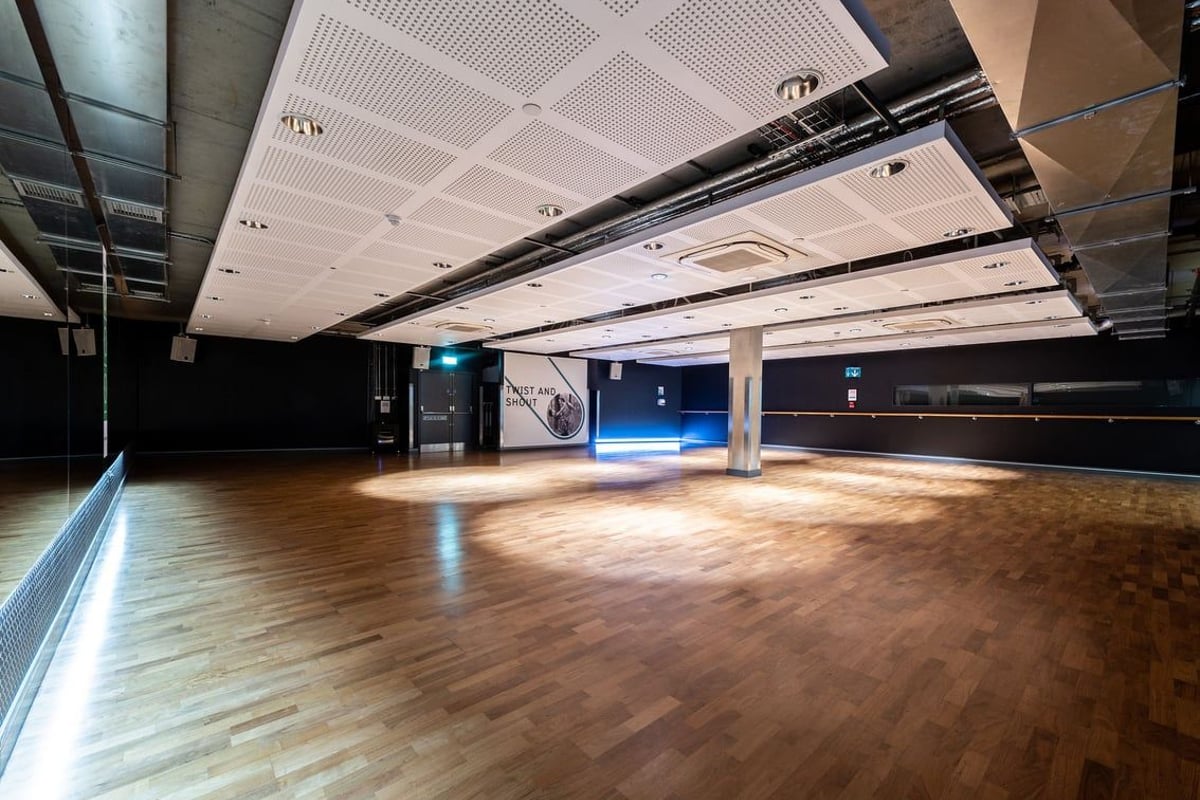 PureGym London Aldgate: Large empty studio space with wooden floors, mirrored walls, and suspended lighting fixtures.