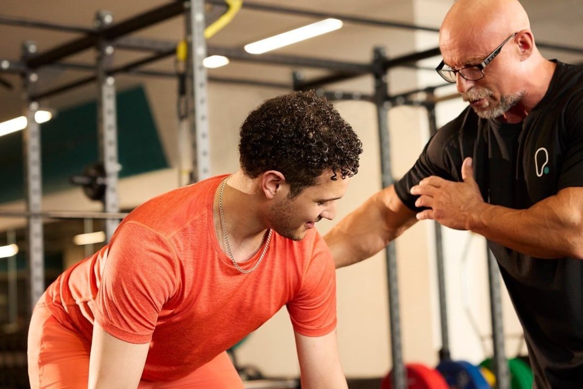 Male gym goer lifting barbell, helped by personal trainer