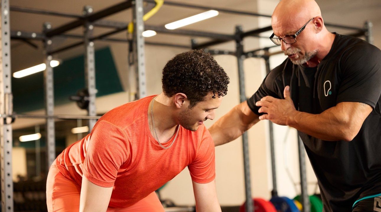 Male gym goer lifting barbell, helped by personal trainer