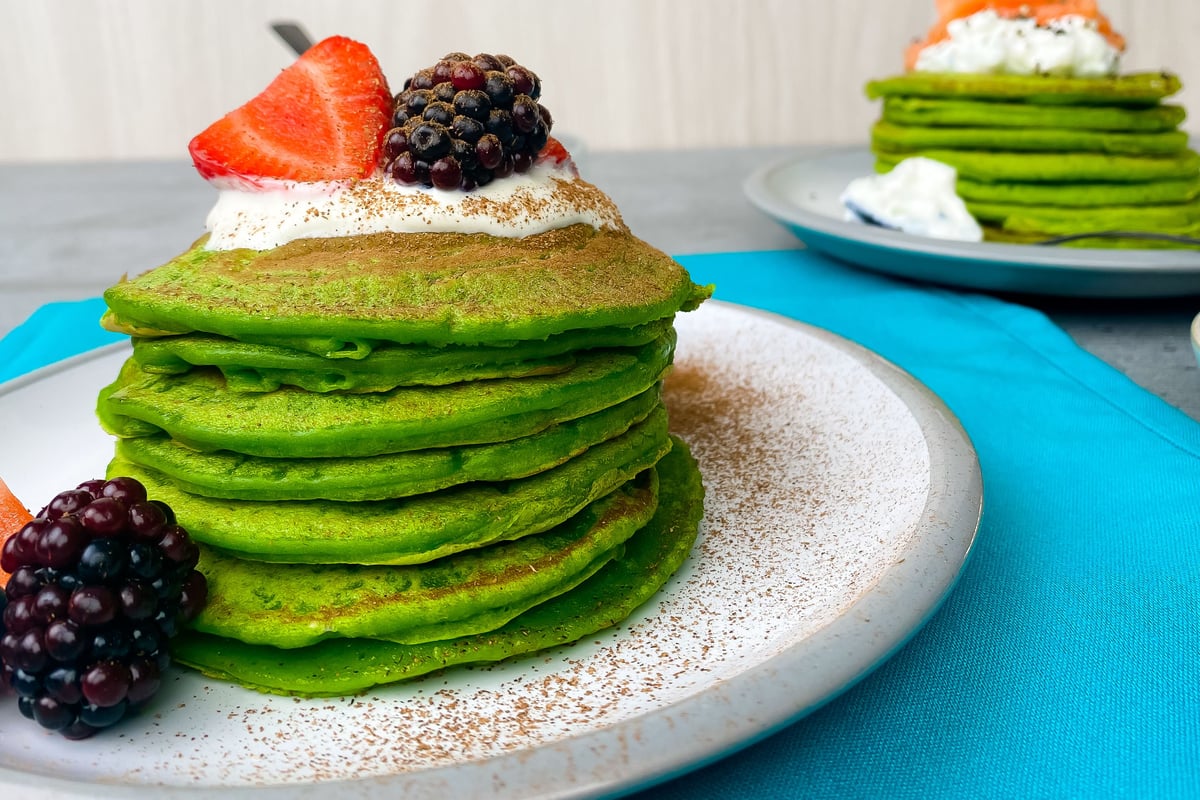 Stack of green pancakes topped with cream, strawberry and blackberry on white plate