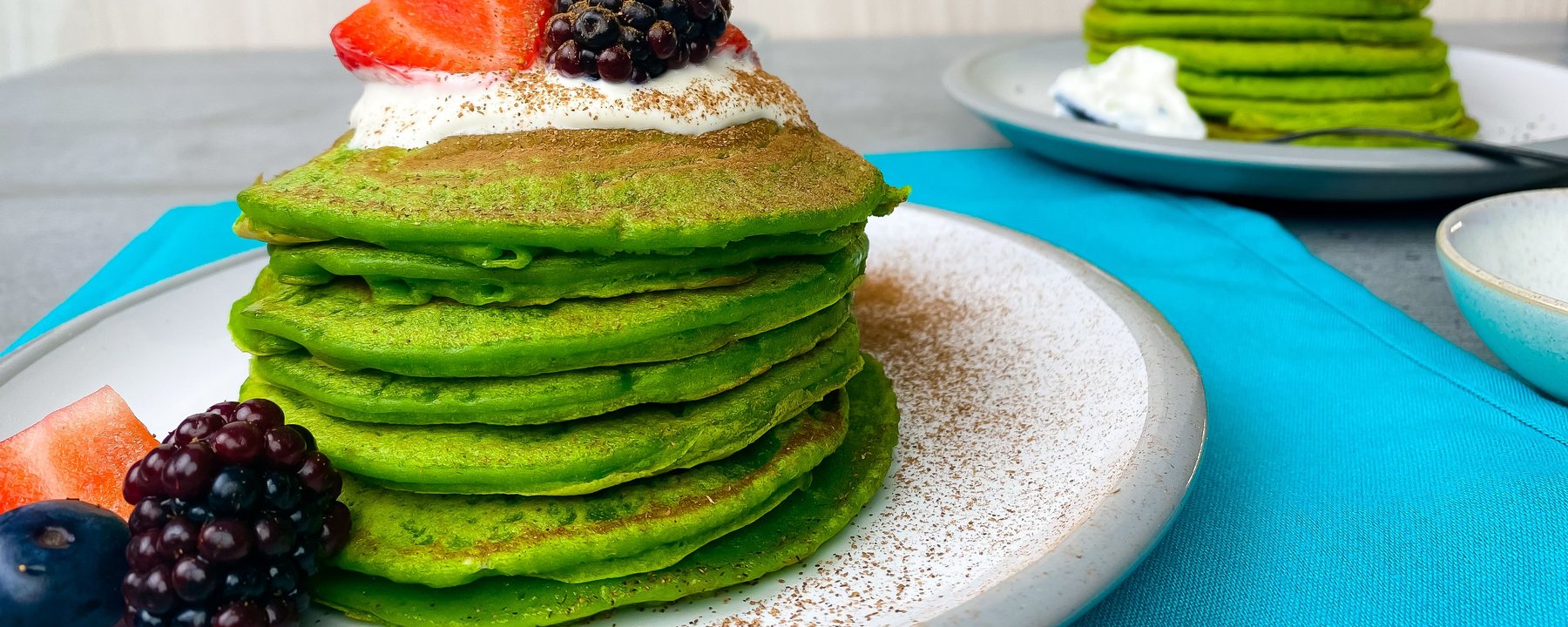 Stack of green pancakes topped with cream, strawberry and blackberry on white plate