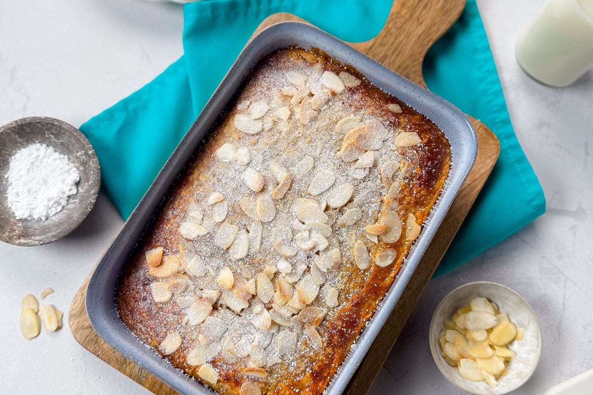 Baked almond cake in a blue tray on a wooden board with powdered sugar and almonds nearby
