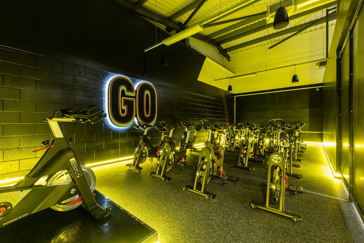 PureGym Ashton Under Lyne: Rows of stationary bikes, neon 'GO' sign, and dimly lit exercise studio.