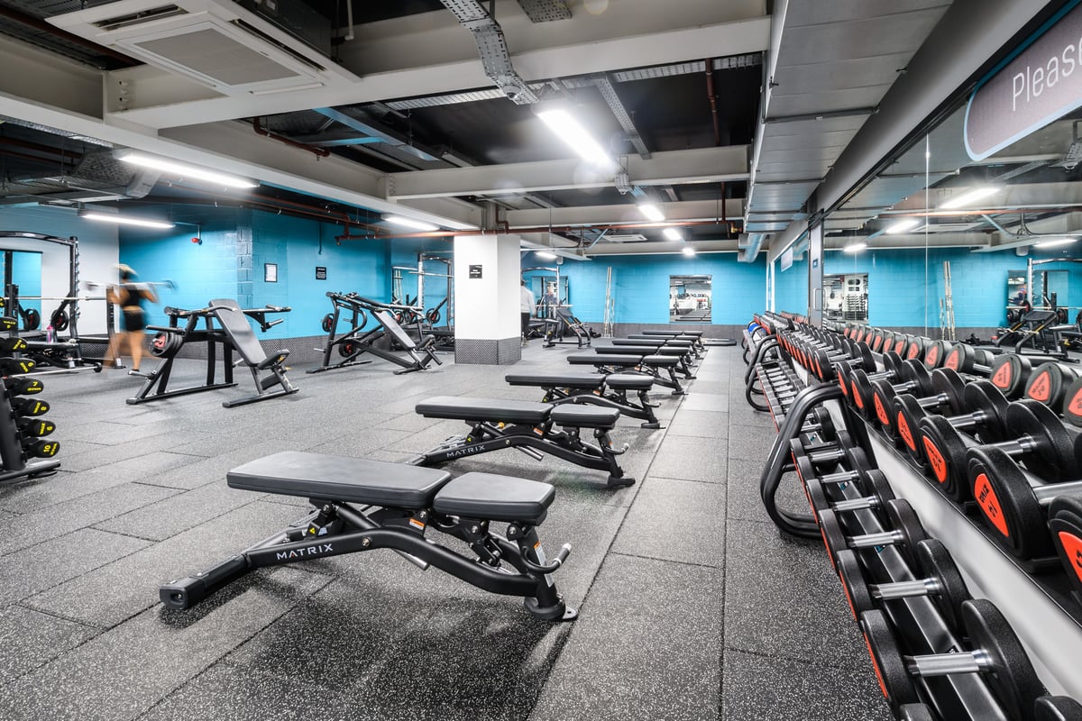 PureGym London East India Dock: Rows of weight benches, machines, free weights, and mirrored walls in a blue-toned gym.