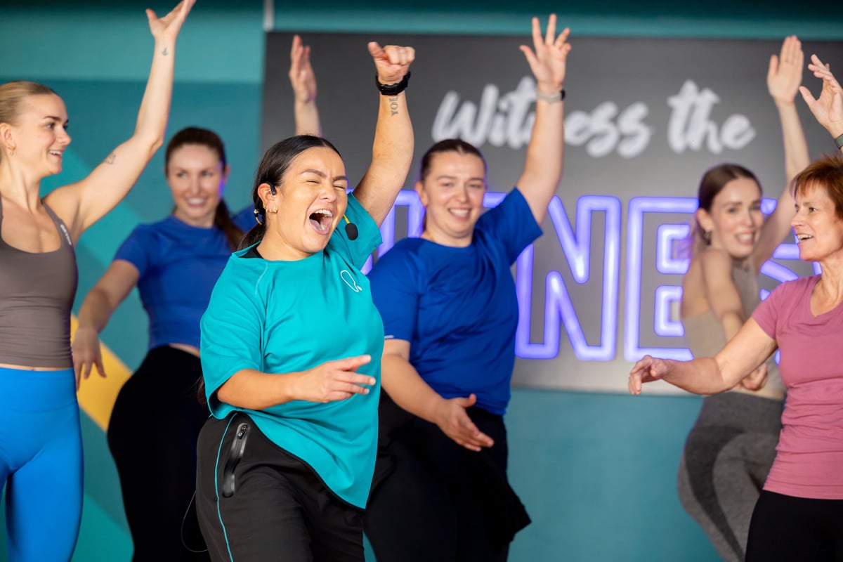 Group of six people smiling and exercising indoors with a neon PURE sign behind them