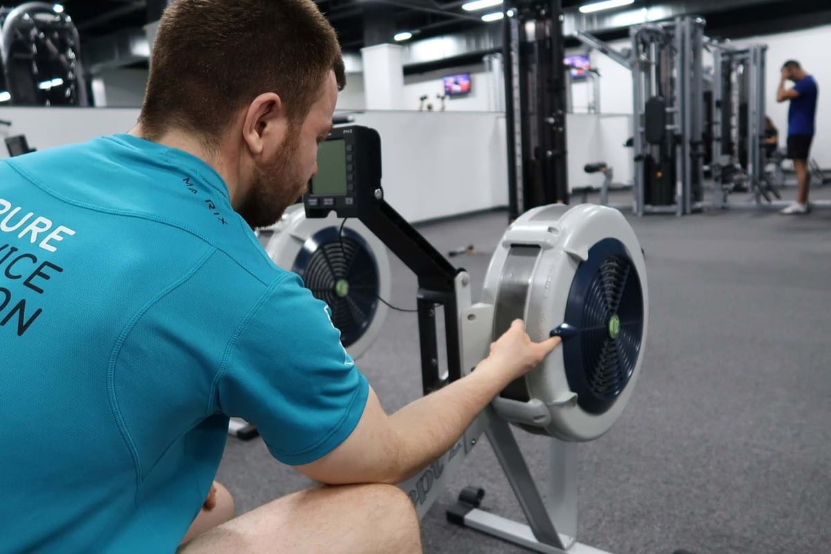 Man adjusting resistance on rowing machine in gym with another person in background