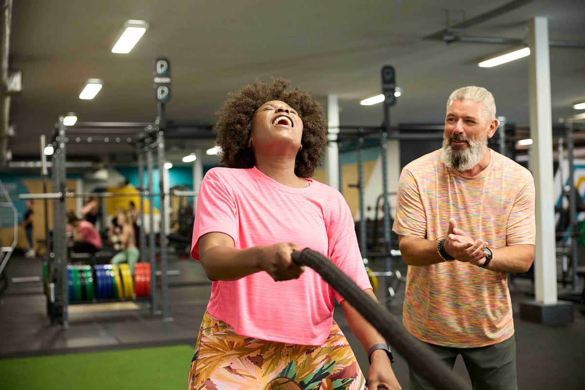 Woman in pink top laughing whilst using battle ropes with man watching in a gym