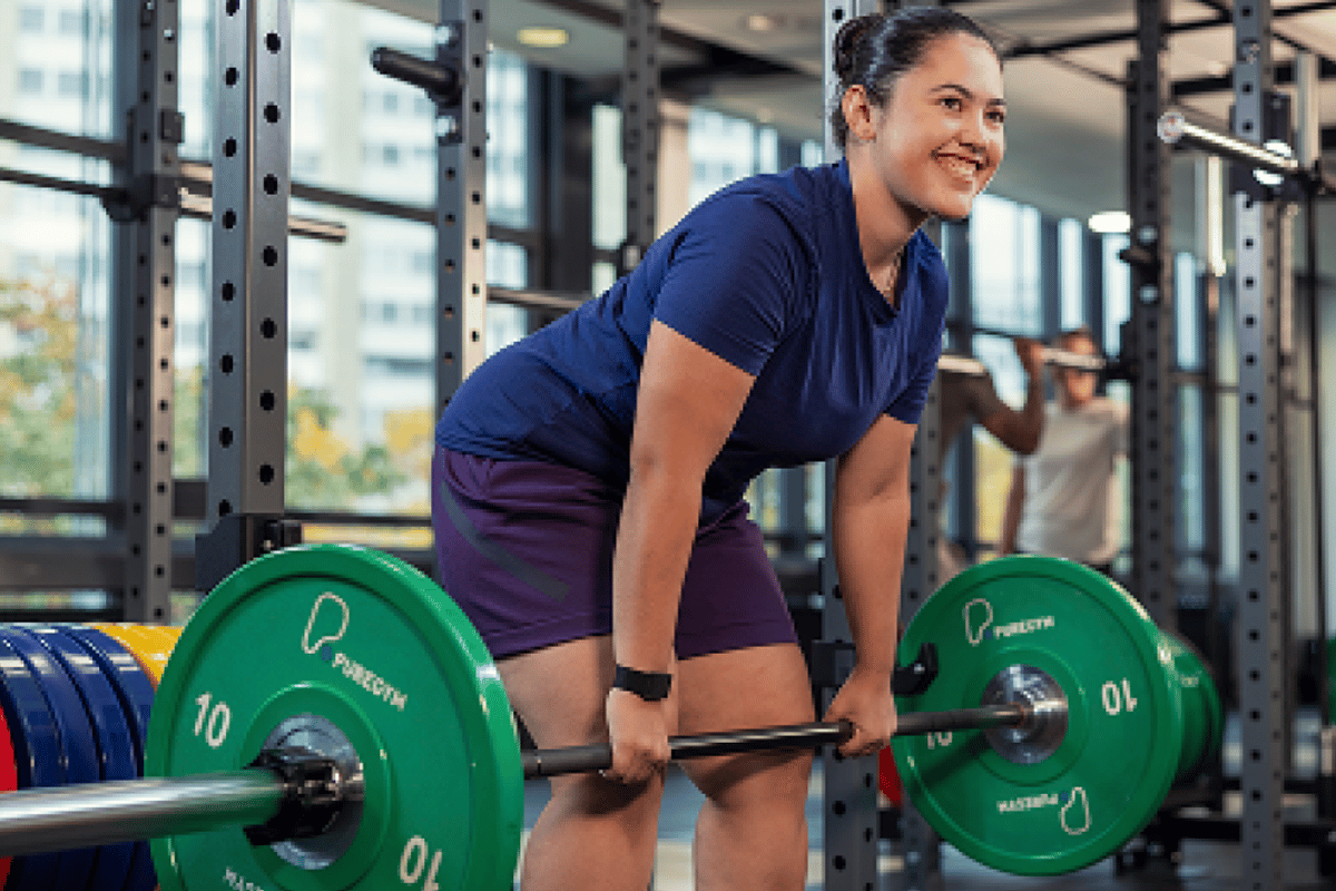 Frau lächelnd beim Kreuzheben mit grüner Langhantel im modernen Fitnessstudio.