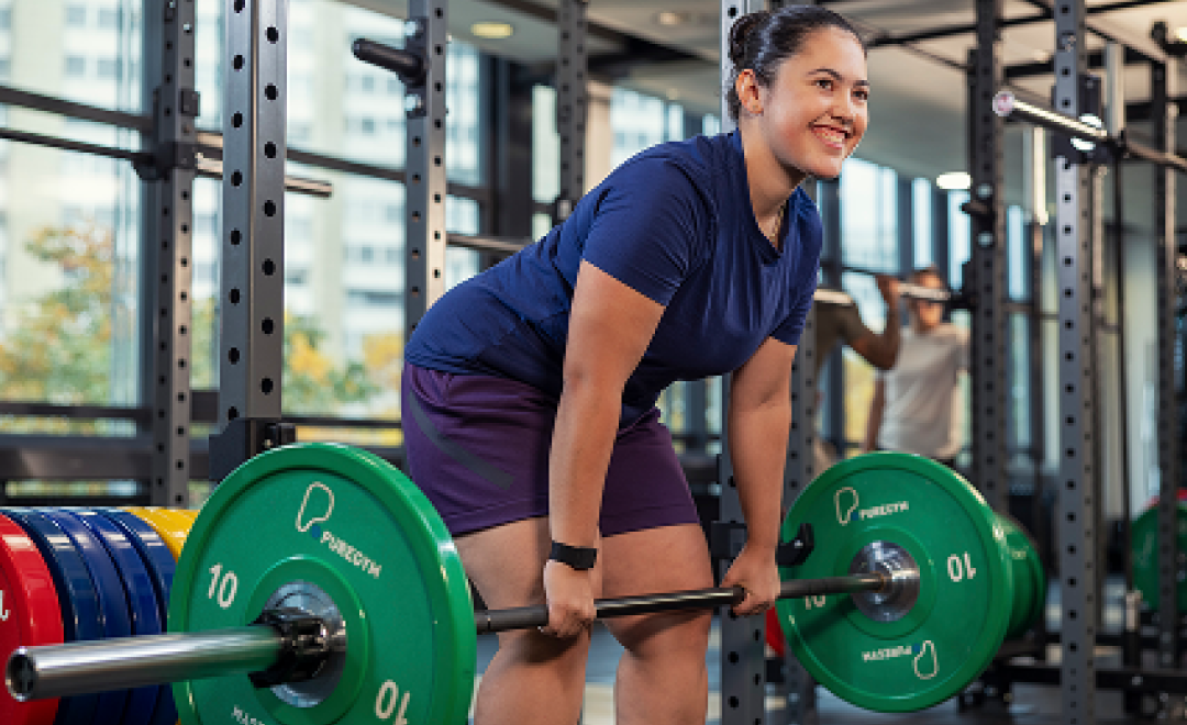 Frau lächelnd beim Kreuzheben mit grüner Langhantel im modernen Fitnessstudio.