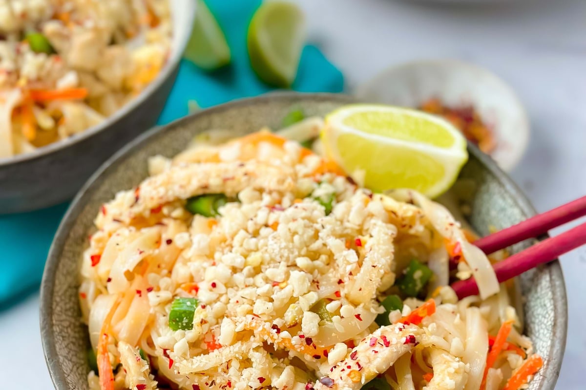 Bowl of pad thai noodles with crushed peanuts, lime wedge, and red chopsticks.