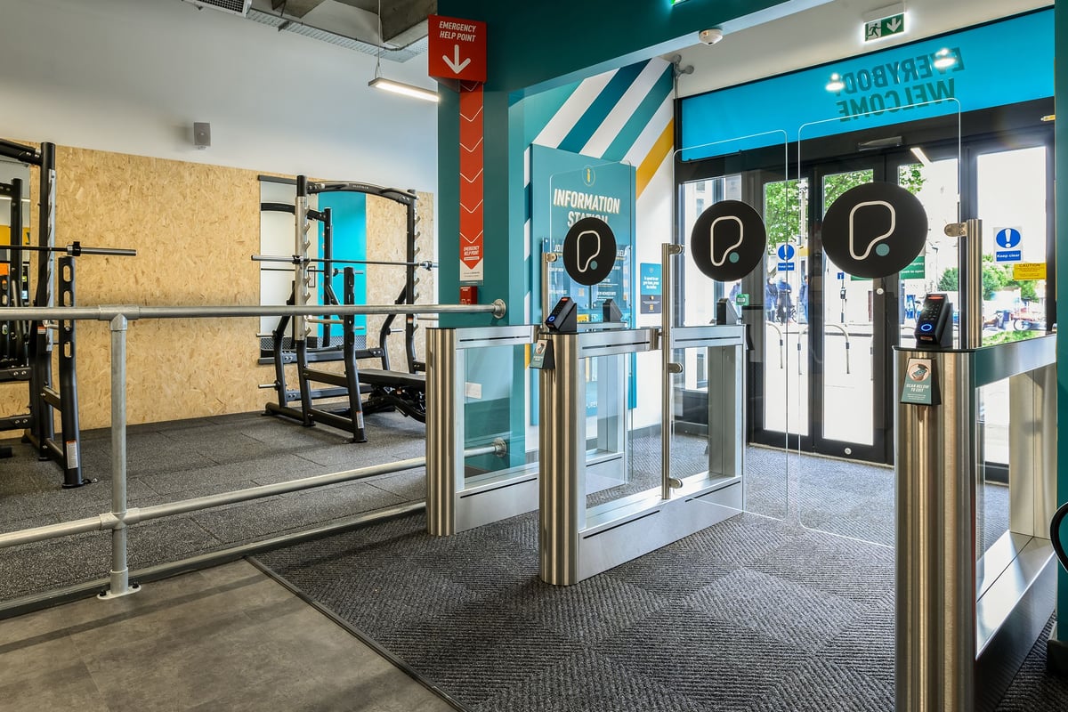 PureGym London Wimbledon: Turnstile entrance gates, digital signage screens, and a waiting area.