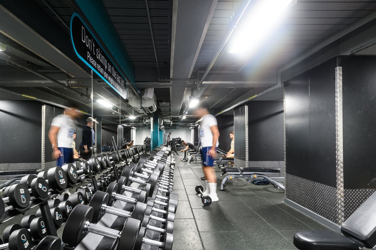 PureGym London Lambeth: Rows of dumbbell racks, weight benches, and rubberized flooring in a dimly lit gym space.