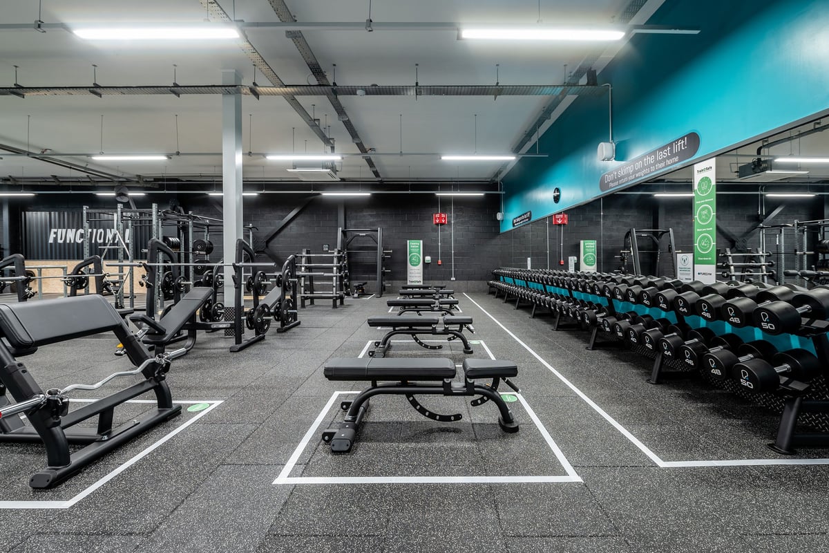 PureGym Birmingham Longbridge: Weight racks, benches, dumbbells, and rubberized floors in a dimly lit gym with teal accents.