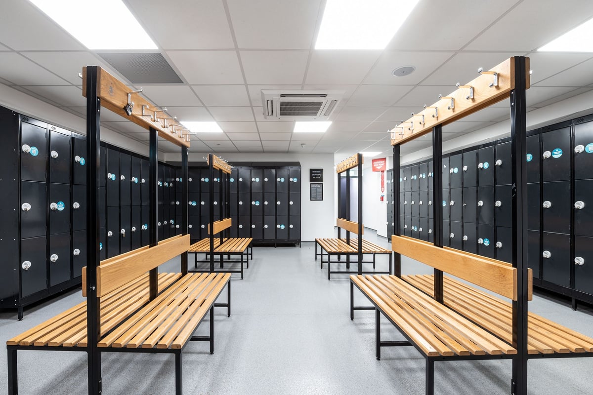 PureGym Sutton Coldfield: Rows of wooden benches and lockers creating an austere changing room environment.