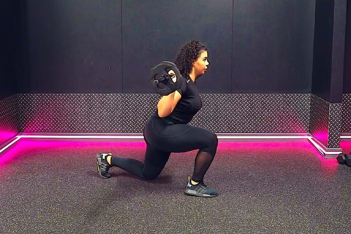 Woman in black workout clothes doing weighted lunges in a gym with pink lights