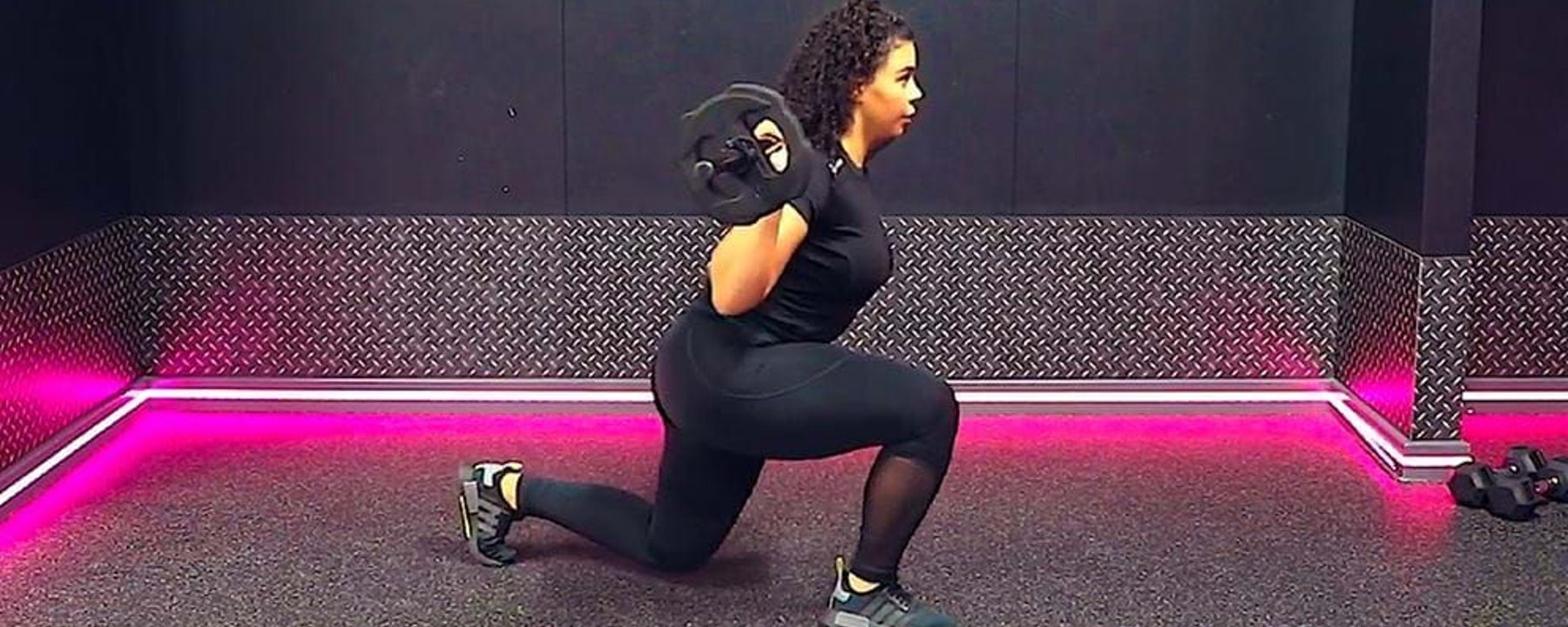 Woman in black workout clothes doing weighted lunges in a gym with pink lights