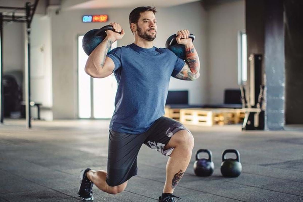 Man in blue shirt doing lunges with kettlebells in a gym with equipment in background