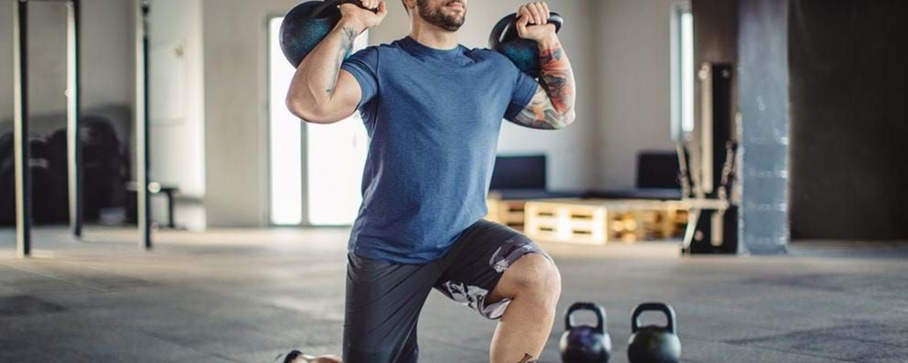 Man in blue shirt doing lunges with kettlebells in a gym with equipment in background