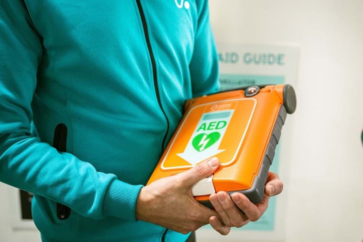 Person in teal jacket holding an orange AED defibrillator device with a green heart symbol