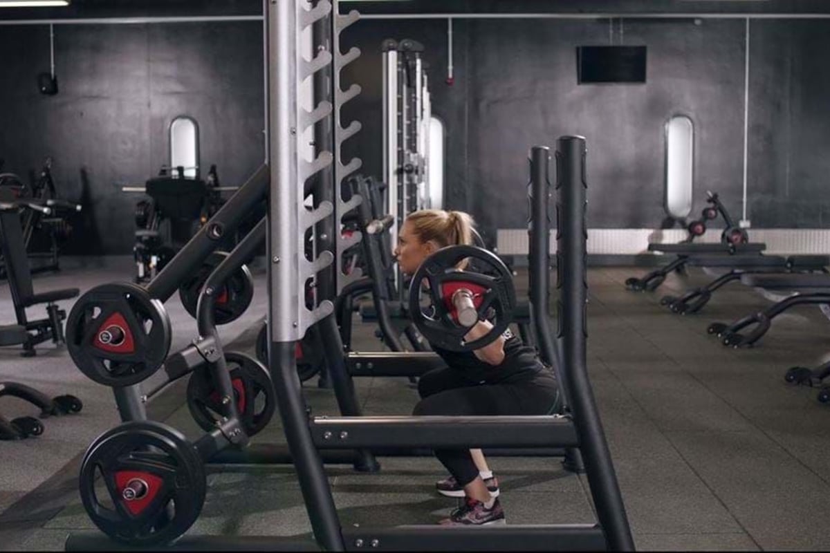 Woman doing barbell squats in a gym with black walls and exercise equipment.