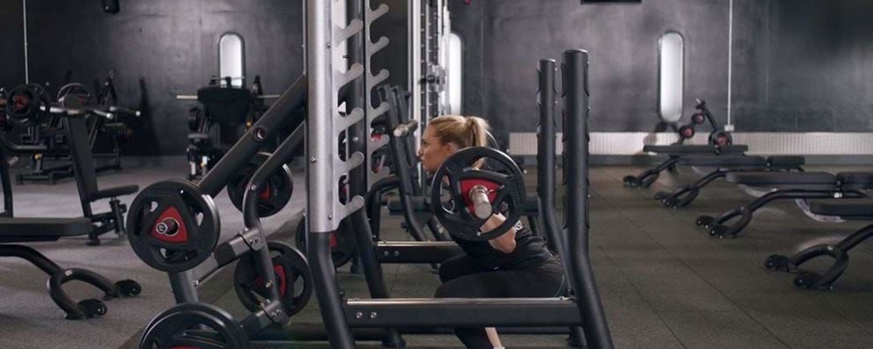 Woman doing barbell squats in a gym with black walls and exercise equipment.
