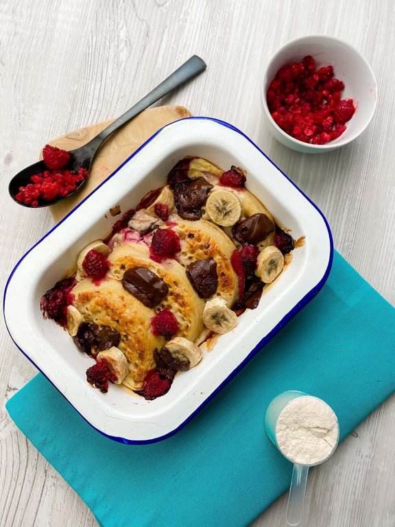 Baked pancakes with chocolate chunks, banana slices, and raspberries in a white tray.