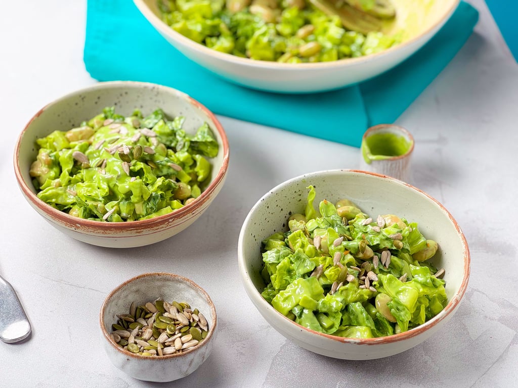 Two bowls of green salad with seeds and a small bowl of mixed seeds on a white surface
