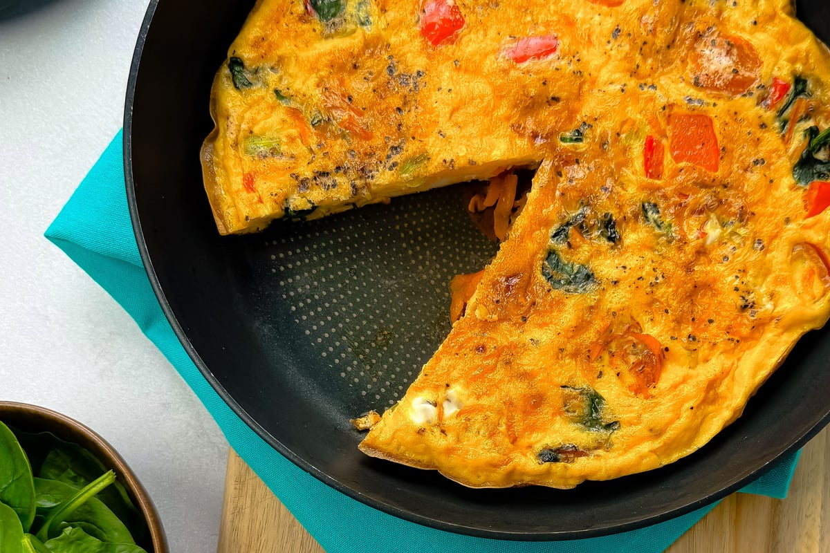 Vegetable omelette with a slice cut out in a frying pan on a blue cloth and wooden board