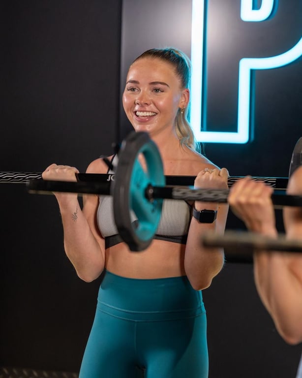 Donna energica sorridente mentre solleva pesi nello studio PureGym moderno con illuminazione al neon