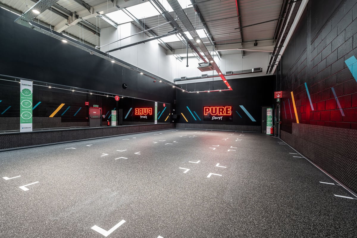 PureGym Stockport North: Bare rubberstudded floor, neon signages, exposed ceiling ducts in dimly lit corridor.