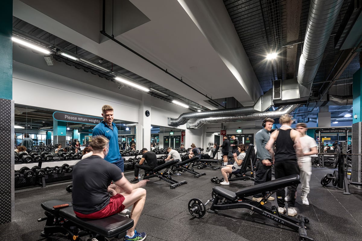PureGym Cardiff Central: Weight benches, rowing machines, weight racks, dumbbells, and people exercising in a well-lit gym.