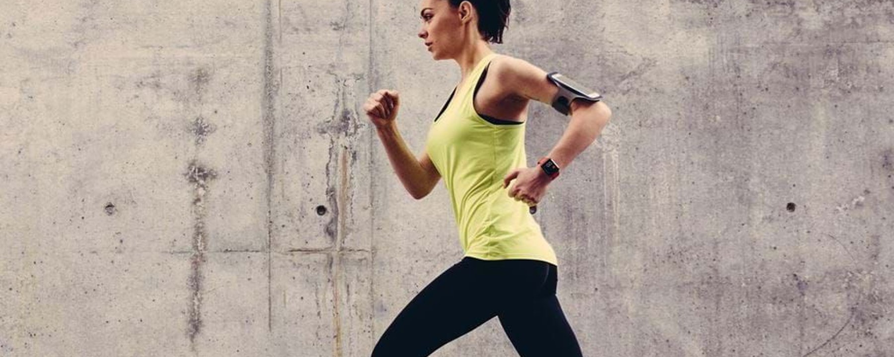 Woman in yellow top and black leggings running against a concrete wall background
