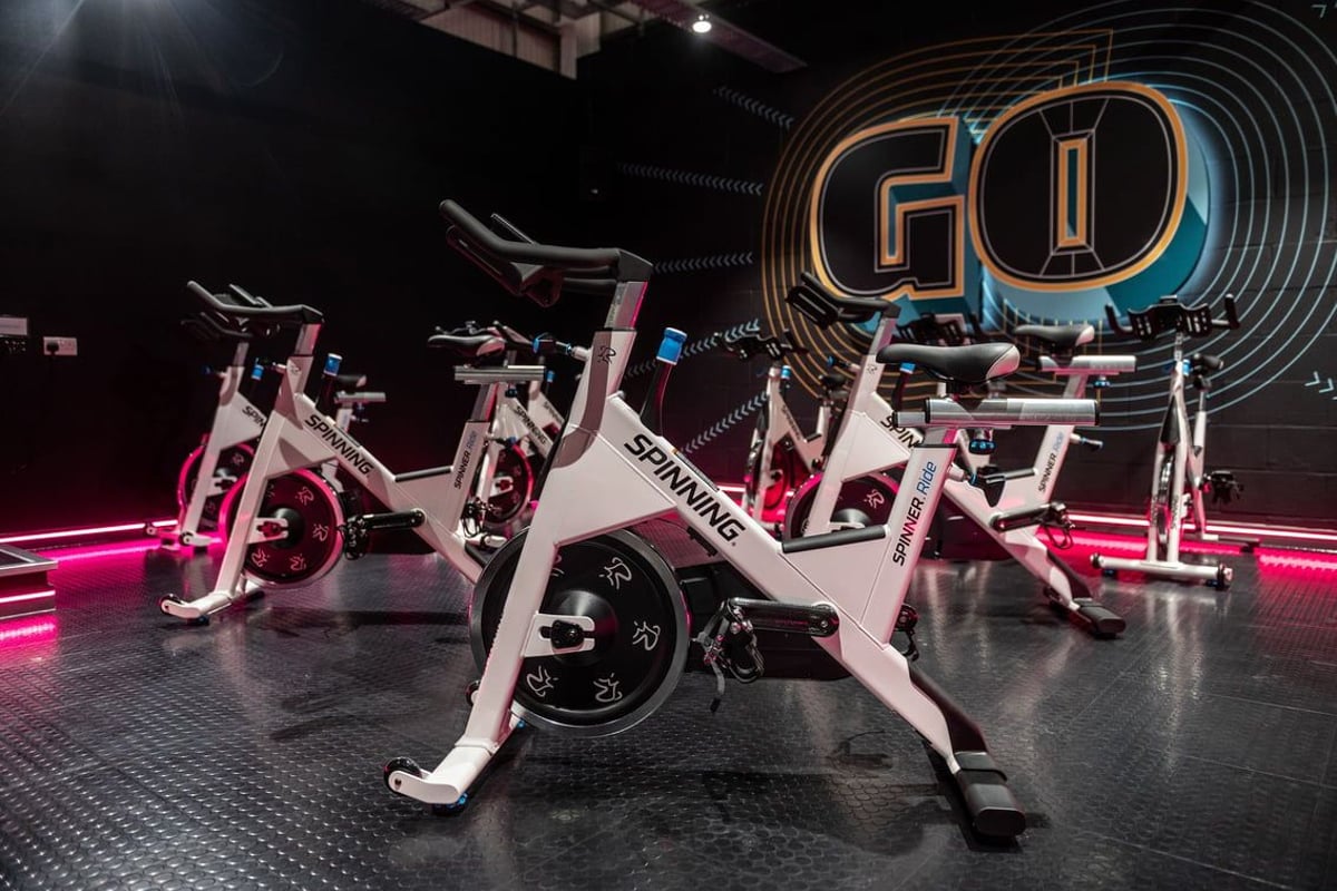 PureGym Swindon Stratton: Rows of stationary bikes, illuminated by neon lights and logos in a dark studio setting.
