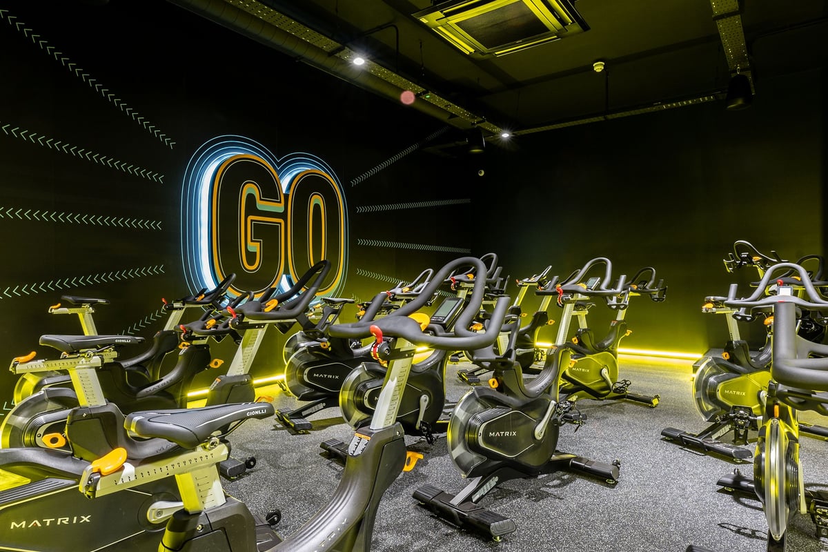 PureGym Kettering: Rows of yellow spin bikes, neon 'Go' sign, black walls with illuminated striped ceiling.