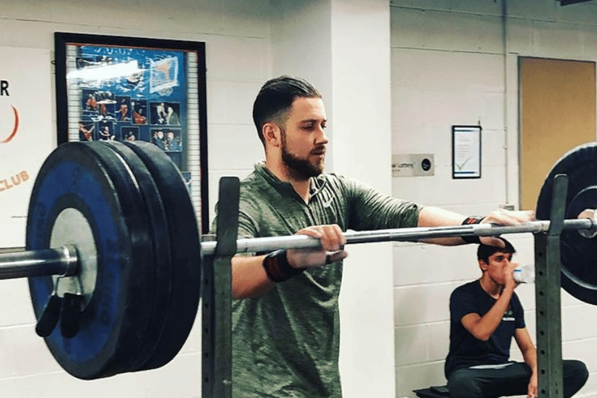 Man adjusting barbell loaded with weights in gym while another drinks water sitting down