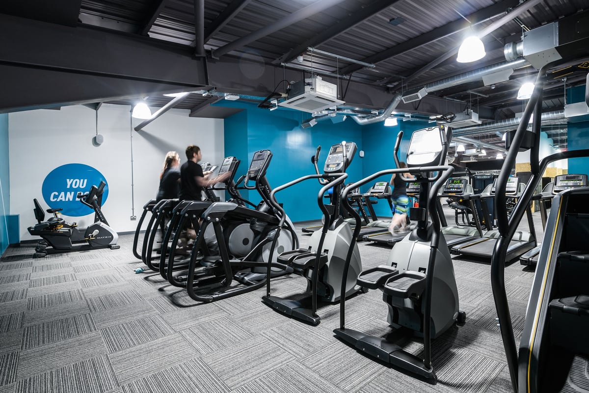 PureGym Hereford: Rows of cardio machines, wheeled arm bikes, and motivational wall art in a bright blue gym space.