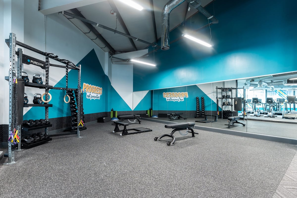 PureGym Loughborough: Functional training rig, squat racks, benches, weight plates, rubberized floors in blue and grey tones.