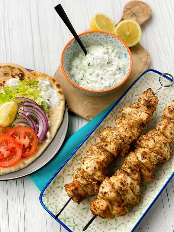 Grilled chicken skewers, flatbread with salad, and a bowl of tzatziki with lemon halves.