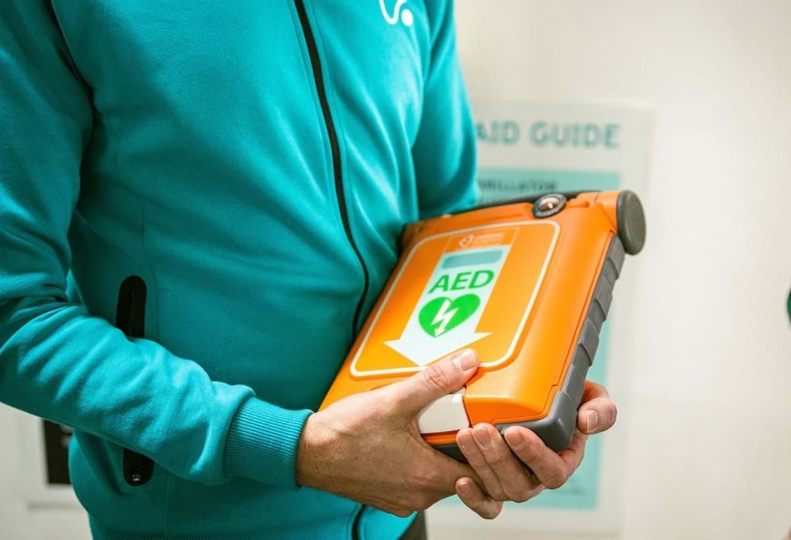 Person in teal jacket holding an orange AED defibrillator device with a green heart symbol