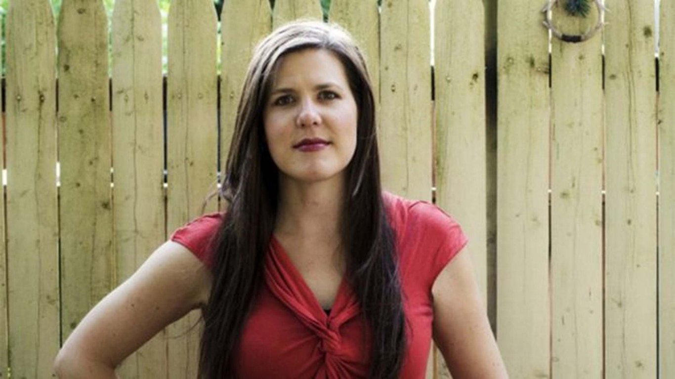 Woman with long dark hair wearing a red top standing in front of a wooden fence