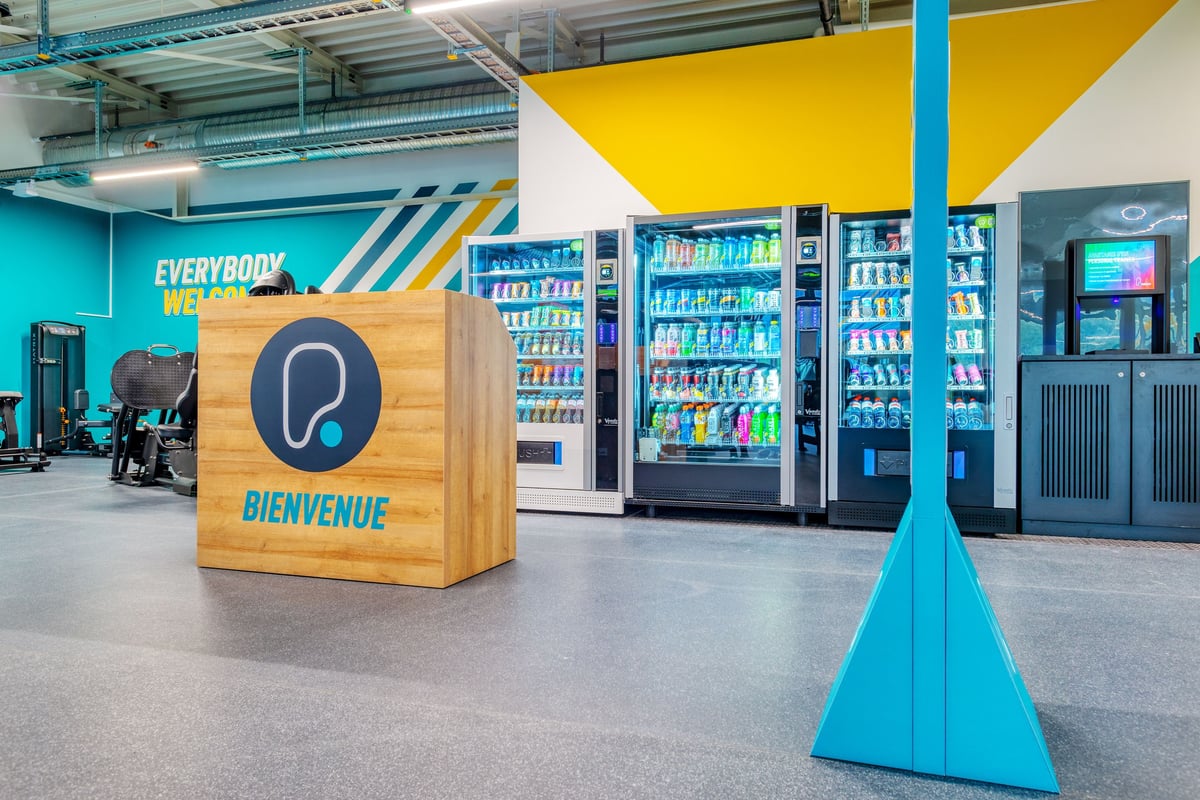 PureGym Martigny: Kühlschränke, Verkaufsautomaten, Sitzwürfel mit Kopfhörer-Logo, blaue Pyramidenskulptur im Fitnessstudio.