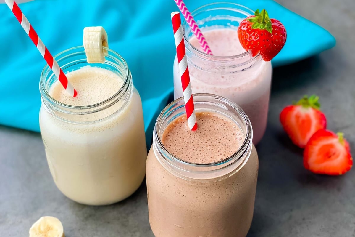 Three jars of banana, strawberry, and chocolate smoothies with straws on grey surface
