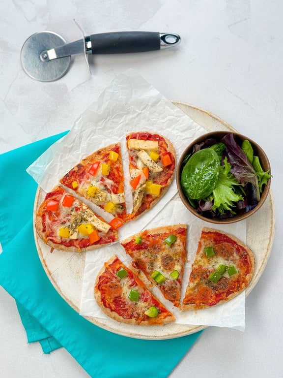 Two small pizzas with vegetables and a bowl of salad on a wooden board with a pizza cutter